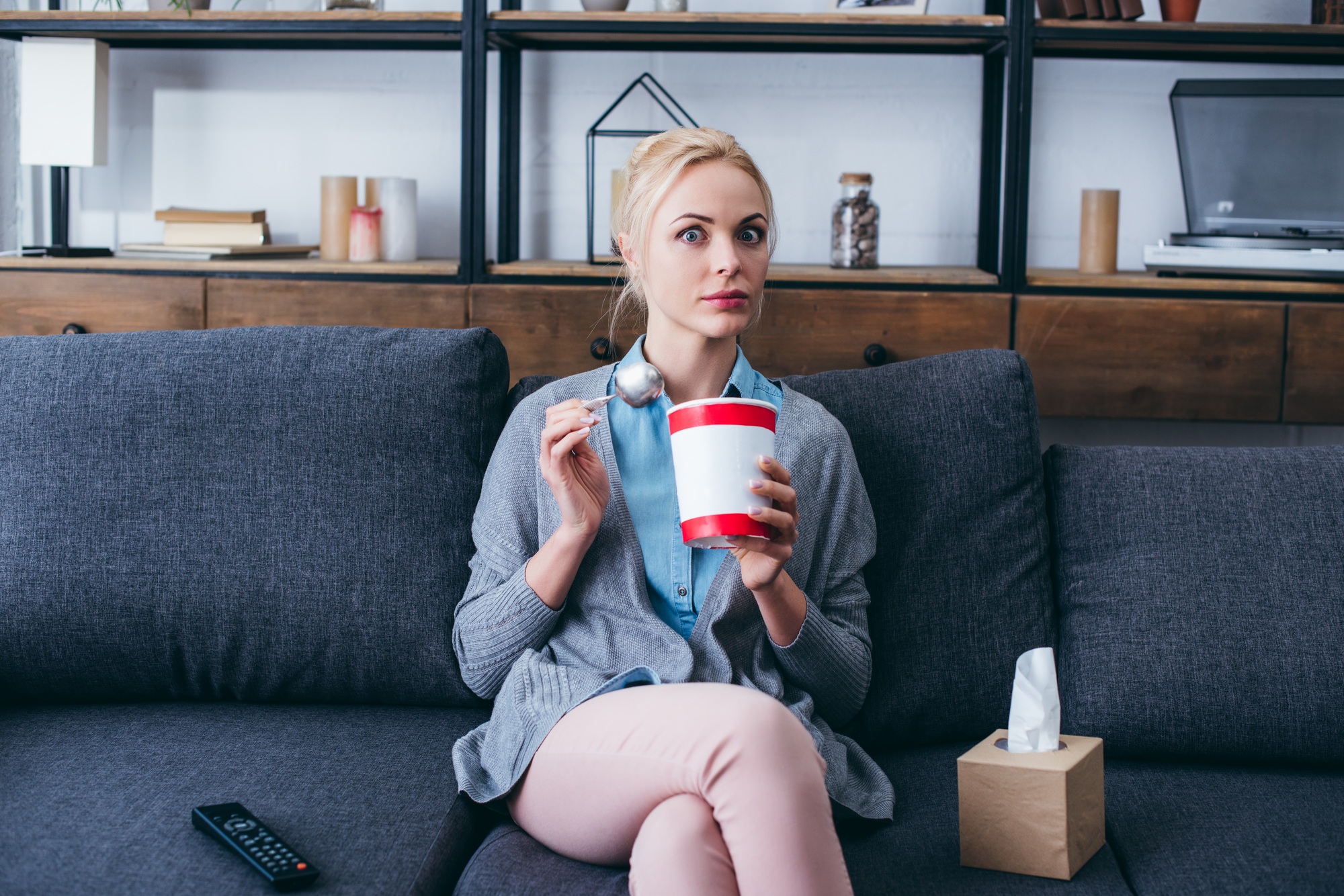 surprised woman eating ice cream and looking at camera while siitng on couch at home alone